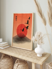 Four black cranes flying across a red sun and stylised mountain against an orange sky, with, in an alternate framed mockup