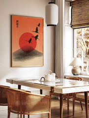 Four black cranes flying across a red sun and stylised mountain against an orange sky, with, in a styled room mockup