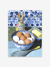 Kitchen Still Life Wall Art showing a bowl of eggs, butter, whisk, and wooden spoons against blue and white tiles.