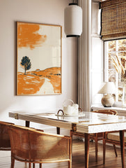 Minimalist landscape art print with orange hillside, dark tree, small house, and abstract orange sky, in a styled room mockup