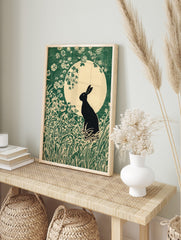 Black rabbit silhouette in a field of green grass and white flowers against a large pale moon, in an alternate framed mockup