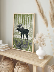 Linocut style print of a black moose standing in water with reflections, surrounded by white, in an alternate framed mockup