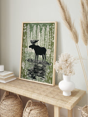Moose silhouette standing in a forest lake with tall trees and birds in a green and white, in an alternate framed mockup