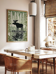 Moose silhouette standing in a forest lake with tall trees and birds in a green and white linocut, in a styled room mockup