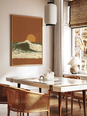 Illustrative ocean wave crashing against a dark teal sea under an orange sunset sky with a large sun, in a styled room mockup