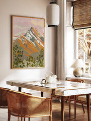 Illustrative mountain landscape print with pink sky, orange peaks, and green foliage foreground, in a styled room mockup