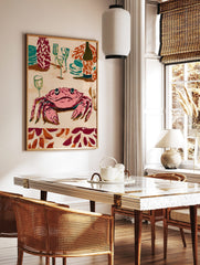 Illustrative wall art print of a pink crab at a dinner table with teal plates, glasses, and a wine, in a styled room mockup