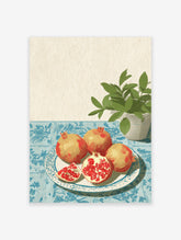 Illustrative pomegranate wall art with whole and sliced pomegranates on a plate, blue tablecloth, and green plant.