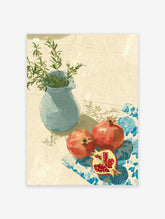 Pomegranate wall art with two red pomegranates, one cut, on a blue patterned cloth next to a blue vase with green leaves.