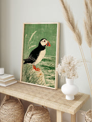 Illustrative print of a puffin bird with a red beak, standing on a rock by water and green, in an alternate framed mockup