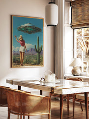 Retro UFO desert wall art featuring a woman waving at a flying saucer above a landscape with cacti and mountains, displayed as the same artwork in a styled poster mockup for home decor