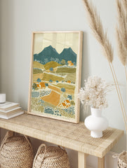 Illustrative rural landscape print with teal mountains, olive green hills, orange fields, and, in an alternate framed mockup
