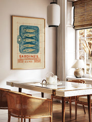 Illustrative linocut-style print of a blue sardine tin with five fish, 'Sardines Dé La Mer' text, in a styled room mockup