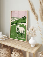 Illustrative print of a white sheep in a green field with rolling hills and pink sky, in a, in an alternate framed mockup