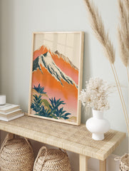 Illustrative mountain landscape art print with snow-capped peaks, orange and pink sky, and, in an alternate framed mockup
