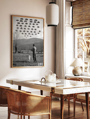 Monochrome print of a man and dog aiming shotgun at pixelated Space Invaders in a rural landscape, in a styled room mockup