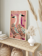 Illustrative wall art of two white spotted geese with green beaks on a textured pink, in an alternate framed mockup