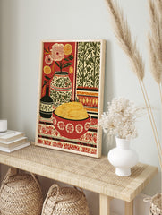 Still life folk art print with a bowl of pasta, flowers in vases, and patterned textiles on a, in an alternate framed mockup