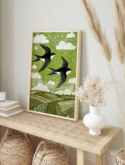 Linocut style print of two black and white swallows flying over green fields and sky with, in an alternate framed mockup