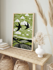 Linocut style print of two black swallows flying over green fields with white clouds, in an alternate framed mockup