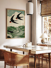Woodcut print of two black and red swallows flying over a green rolling landscape with fields and, in a styled room mockup