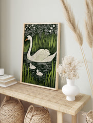White swan and two cygnets swimming in dark water, surrounded by green reeds and white, in an alternate framed mockup