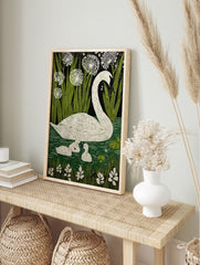 White swan and two cygnets swimming in green water with reeds and white flowers against a, in an alternate framed mockup