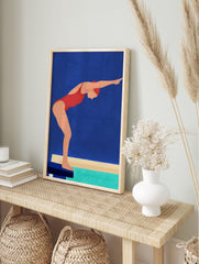 Illustrative wall art of a swimmer in a red swimsuit and cap, poised on a starting block above turquoise water, against a blue background, displayed as the same artwork in an alternate framed wall art mockup
