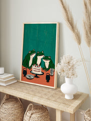 Illustrative wall art featuring three green frogs looking at a white cake with red berries on, in an alternate framed mockup