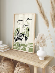 Minimalist illustration of three herons with black outlines and olive green bodies standing, in an alternate framed mockup