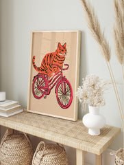 Orange tiger-striped cat riding a pink bicycle illustration on a textured beige background, in an alternate framed mockup