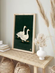 Illustrative print of two white geese with orange beaks walking on a dark green background, in an alternate framed mockup