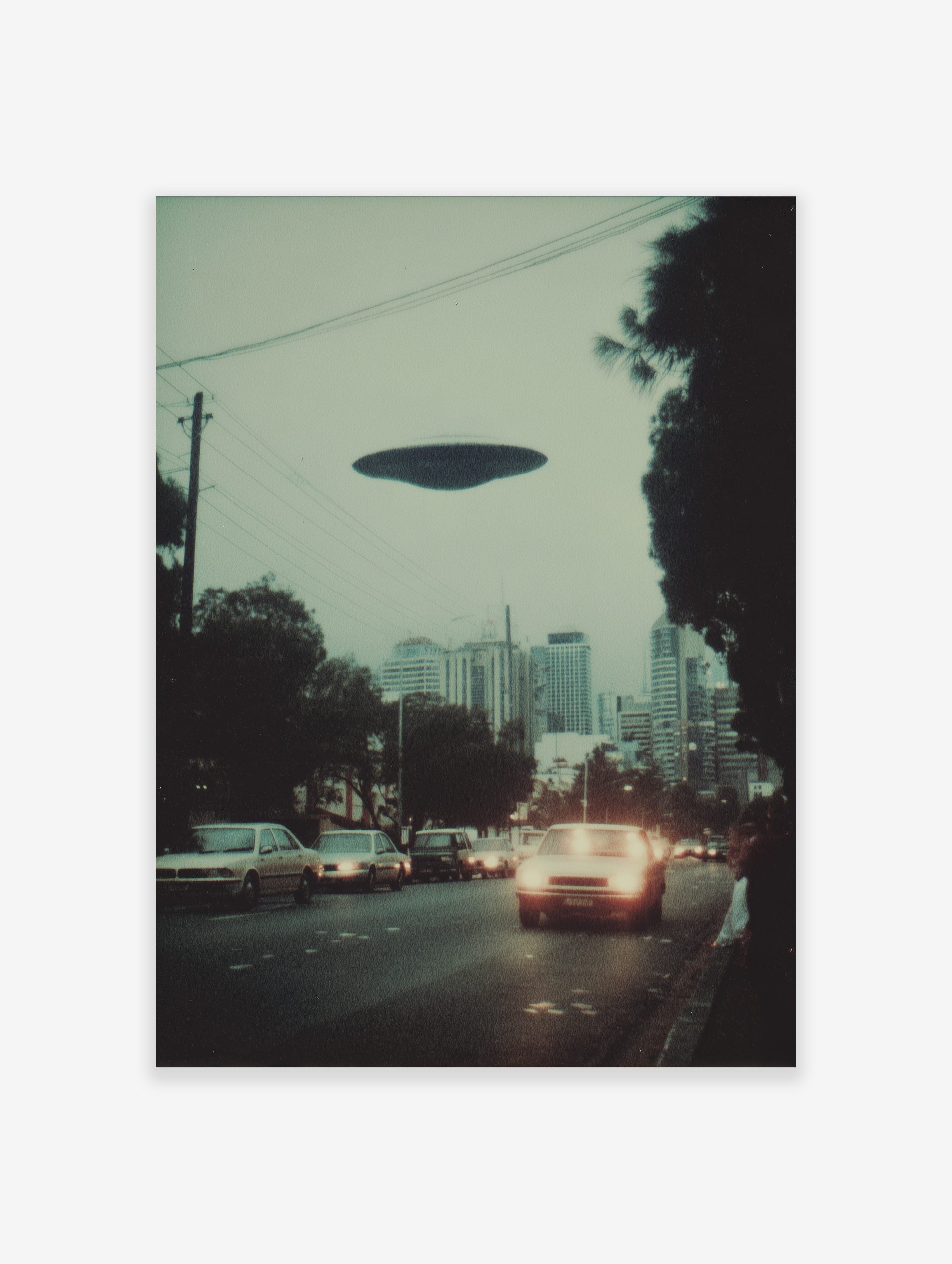 Photographic print of a dark disc-shaped UFO hovering over a city street with cars and buildings at dusk