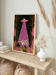 Whimsical illustrative wall art of a black and white cow being abducted by a purple UFO beam, in an alternate framed mockup