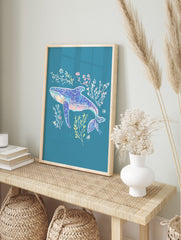 Illustrative blue whale art print with pink, yellow, and white seaweed on a teal background, in an alternate framed mockup