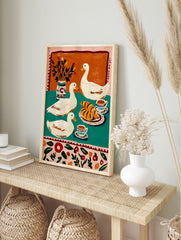 Illustrative print of three white ducks with croissants, coffee, and a vase on a teal and, in an alternate framed mockup