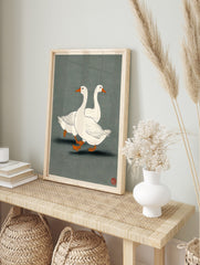 Illustrative print of two white geese with orange beaks and feet walking on a dark grey-green, in an alternate framed mockup