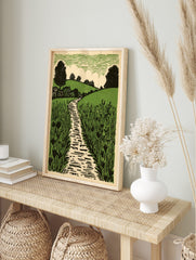 Linocut-style art print of a winding path through green fields with trees and a textured sky, in an alternate framed mockup