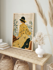 Illustrative art print of a woman in yellow dress and black hat, looking at green leaves with, in an alternate framed mockup