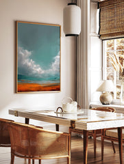 Abstract landscape art print with teal-grey sky, white clouds, and vibrant orange-blue ground, in a styled room mockup