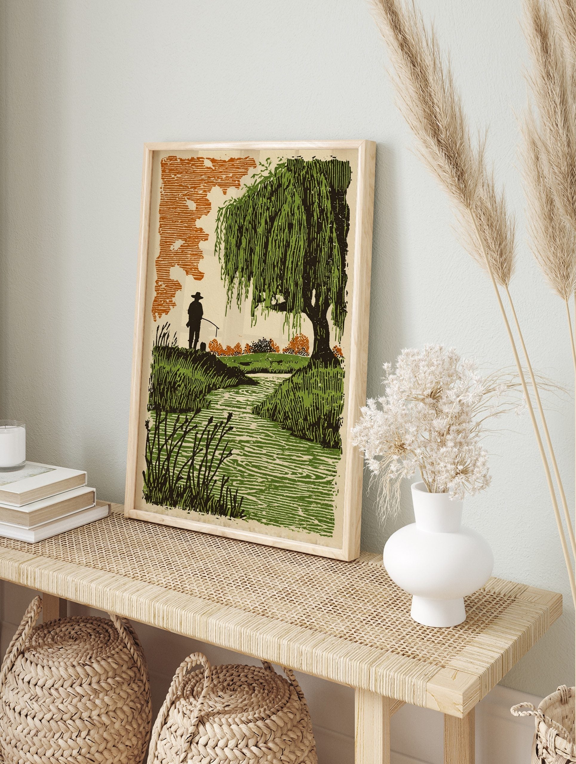 Lino-cut style print of an angler fishing by a green stream with weeping willow tree and, in an alternate framed mockup