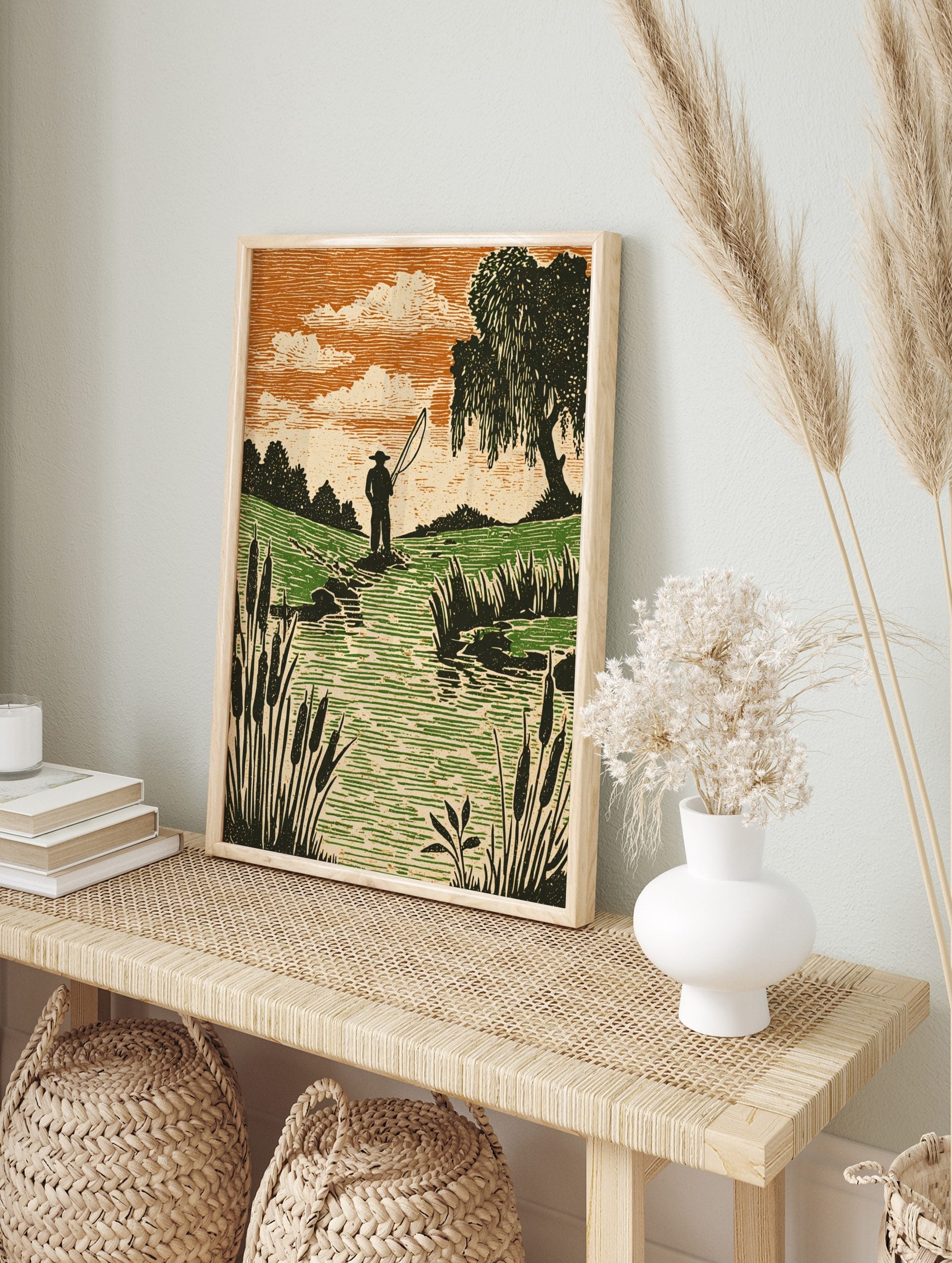 Woodcut style print of an angler fishing in a stream with green fields, orange sky, and, in an alternate framed mockup