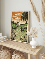 Woodcut style print of an angler fishing in a stream with green fields, orange sky, and, in an alternate framed mockup