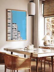 Minimalist architectural wall art of modern white and pink apartment buildings against a clear blue sky, displayed as the same artwork in a styled poster mockup for home decor