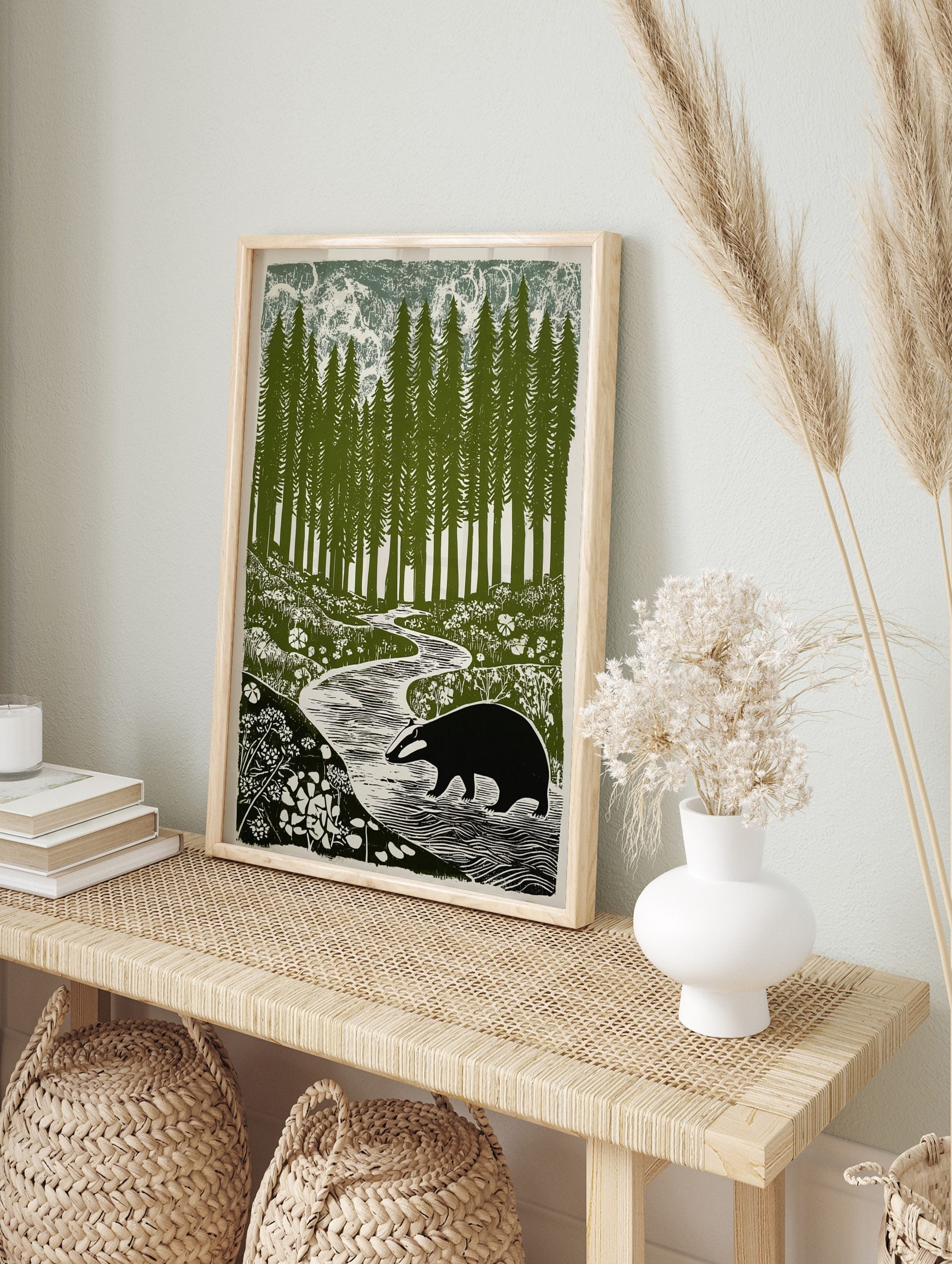 Linocut-style print of a badger walking by a winding river in a forest of tall pine trees and, in an alternate framed mockup