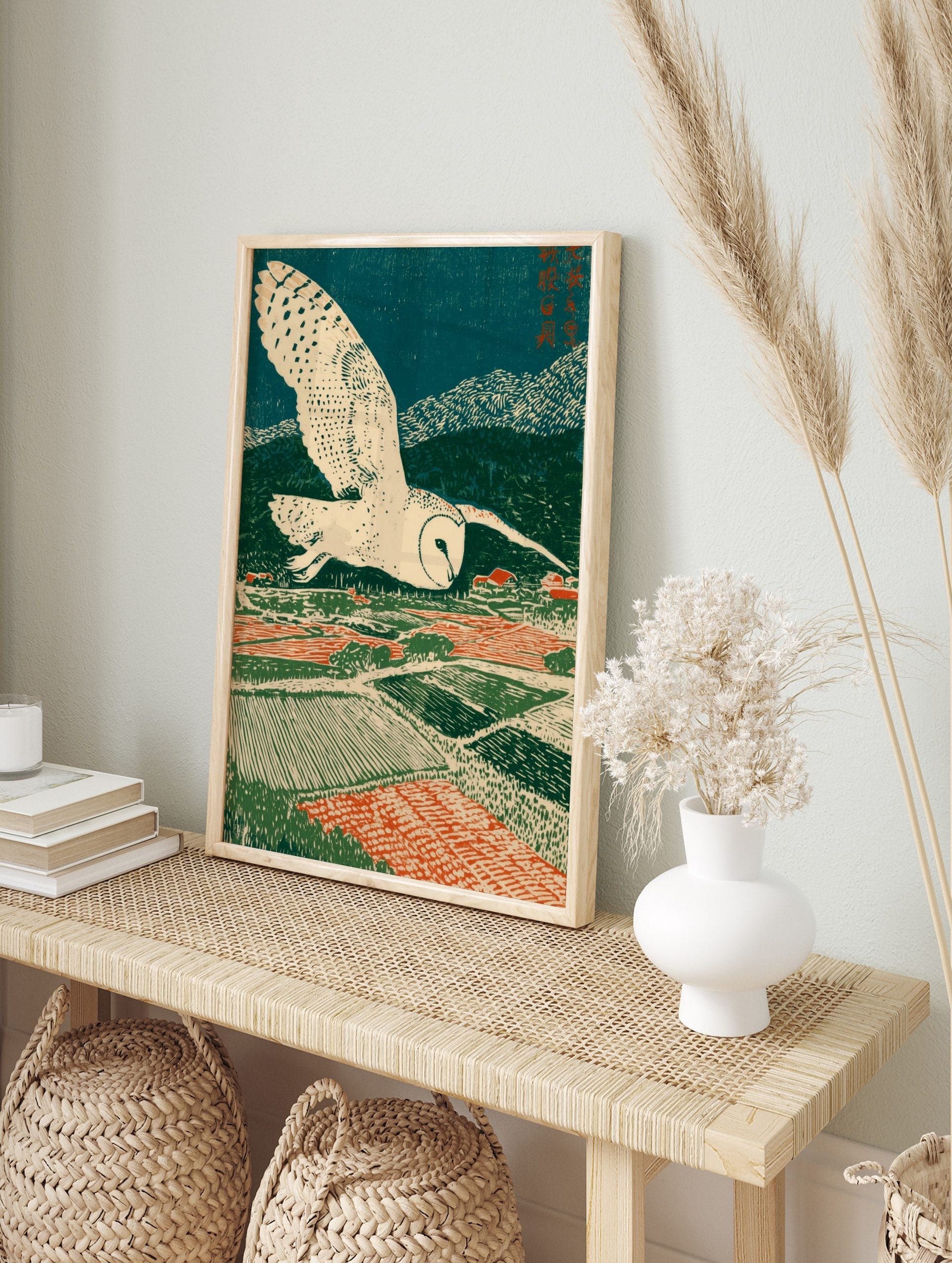 Stylised barn owl flying over patterned green and red fields with a village under a dark blue, in an alternate framed mockup