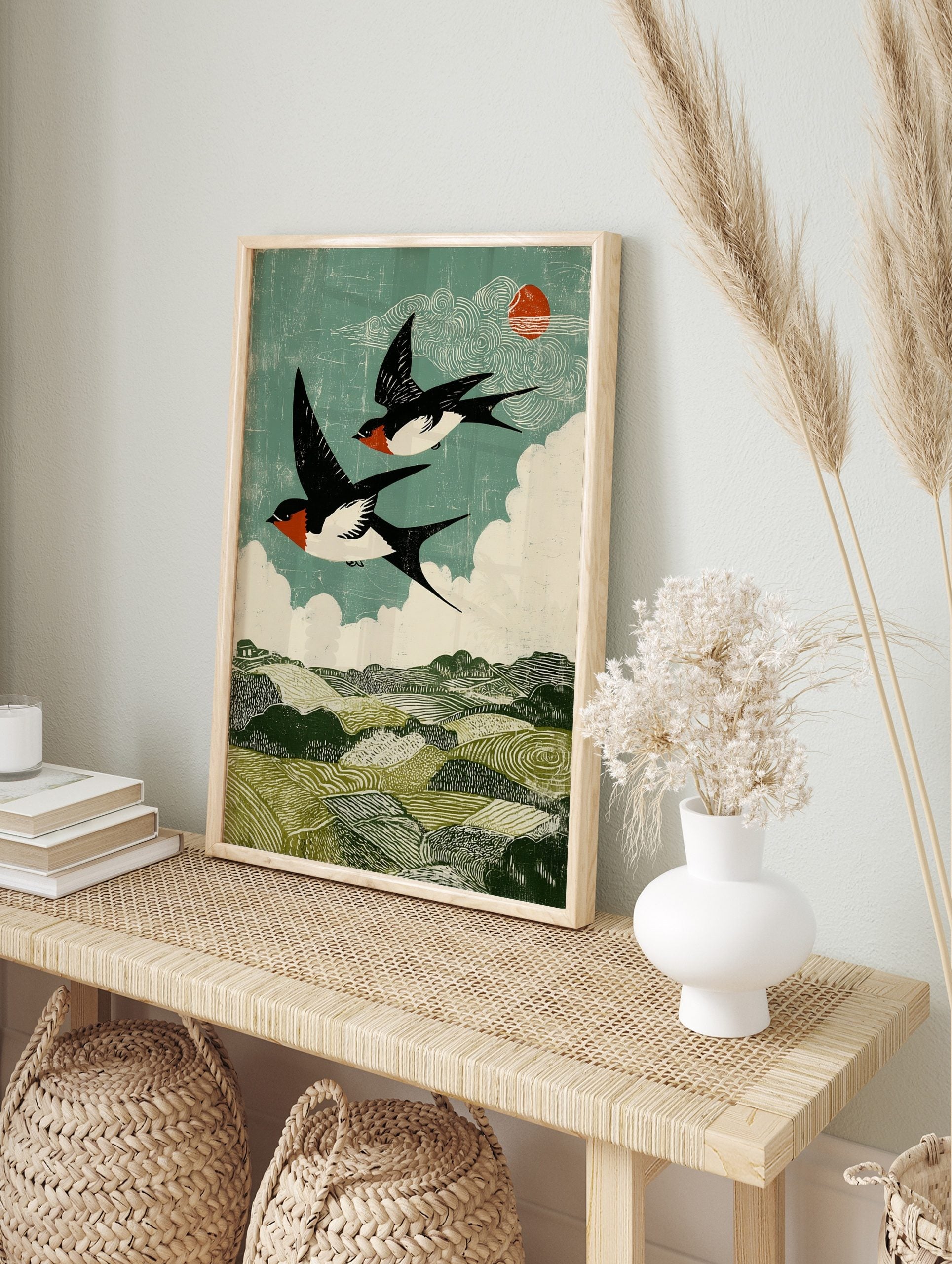 Two barn swallows flying over green hills under a teal sky with white clouds and red sun in, in an alternate framed mockup