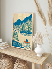 Illustrative print of a blue bear in an orange canoe on a lake with pine trees and blue, in an alternate framed mockup