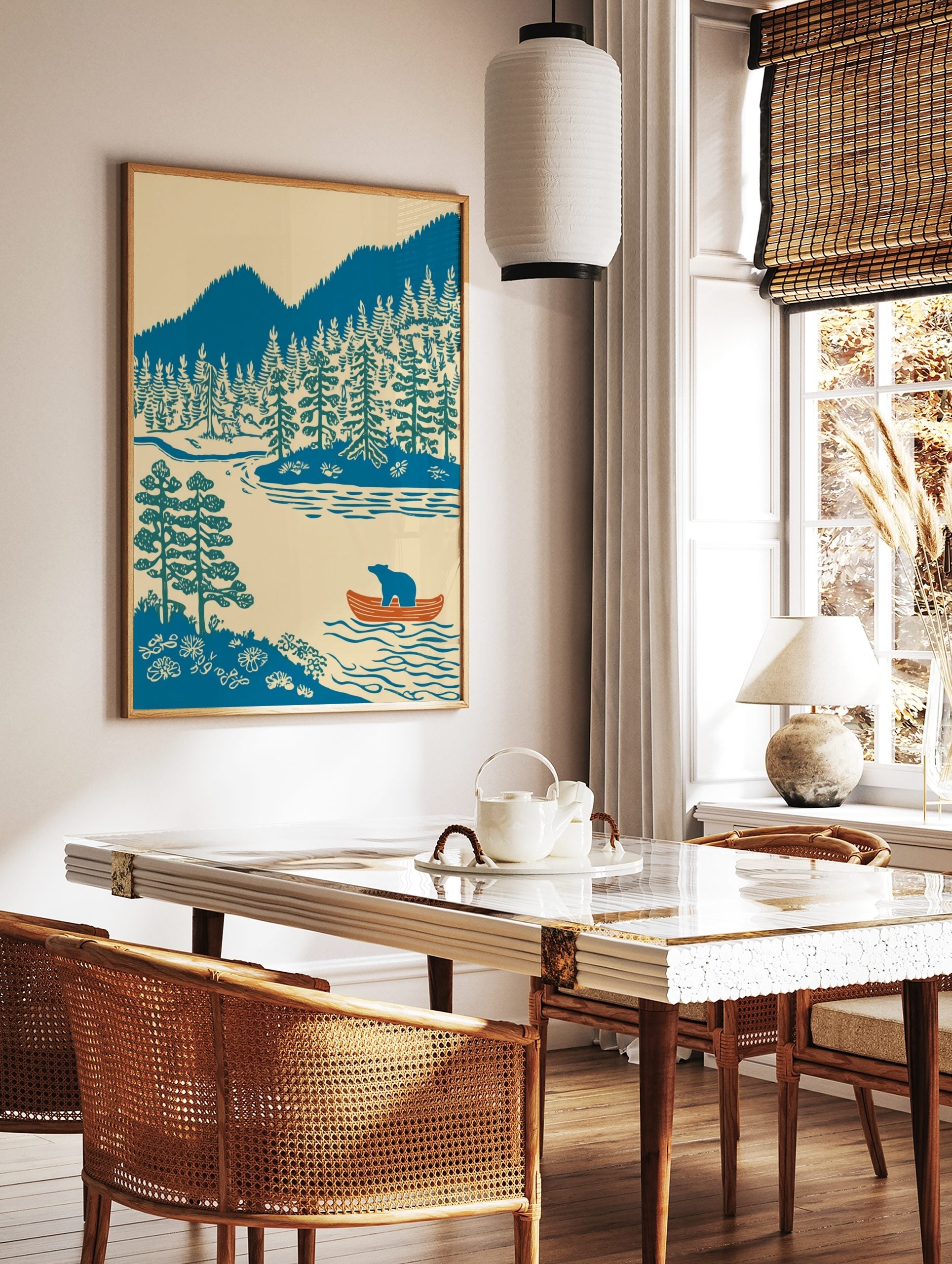 Illustrative print of a blue bear in an orange canoe on a lake with pine trees and blue mountains, in a styled room mockup