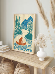 Brown bear paddling orange canoe on blue river with teal trees and blue mountains in, in an alternate framed mockup